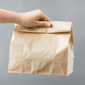 recycling and ecology concept - hand holding disposable brown takeaway food in paper bag with lunch on table